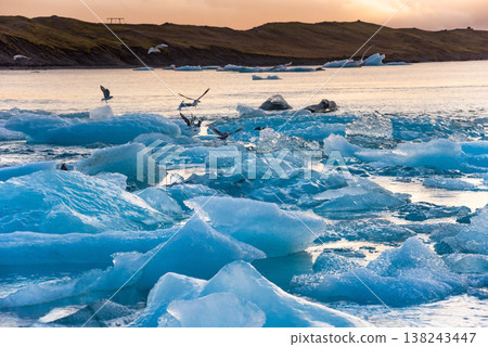 Icebergs float in Iceland's glacier lagoon, reflecting the warm colors of sunset. Birds fly above, adding life to the tranquil scene. This location is a must-see for nature enthusiasts. 138243447