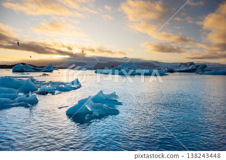 Icebergs float in Iceland's glacier lagoon, reflecting the warm colors of sunset. Birds fly above, adding life to the tranquil scene. This location is a must-see for nature enthusiasts. 138243448