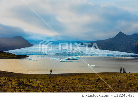 Visitors admire the breathtaking scenery of Fjallsarlon lagoon in Iceland, where majestic glaciers meet floating icebergs under a cloudy sky 138243455