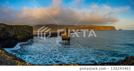 Kirkjufjara beach showcases black sand and unique rock formations under a vibrant sunset. The coastline features gentle waves and scenic mountains in the background. 138243461