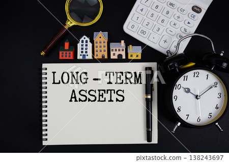 Notepad with the phrase "Long - Term Assets" surrounded by a pen, calculator, magnifying glass, clock, and house model 138243697