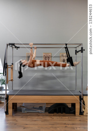 Woman performing extended leg pendulum pose on pilates Cadillac machine in modern studio. Extended, pendulum pose, Pilates Cadillac, modern studio, active. 138244653