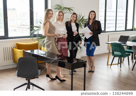 Four professional businesswomen standing together in a modern brightly lit office discussing strategy and holding documents, representing teamwork, ambition, leadership, and corporate success Four professional businesswomen standing together in a modern brightly lit office discussing strategy and holding documents, representing teamwork, ambition, leadership, and corporate success 138244899