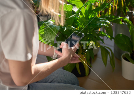 Woman holding smartphone and touching yellow leaf of peace lily in pot indoors Woman holding smartphone and touching yellow leaf of peace lily in pot indoors 138247623