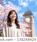 A spring portrait of a woman smiling and looking up against the backdrop of a clock tower, set against a blue sky and cherry blossoms in full bloom. 138248429