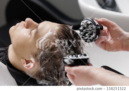 Hairdresser uses shampoo and a scalp brush to wash a boy hair at a salon sink. Close-up of scalp care, hygiene routine, and grooming service process. Hairdresser uses shampoo and a scalp brush to wash a boy hair at a salon sink. Close-up of scalp care, hygiene routine, and grooming service process. 138251522