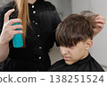 Hairdresser applies styling spray to a boy hair after a haircut. Close-up of hairstyling process, grooming routine, and finishing service in salon. 138251524