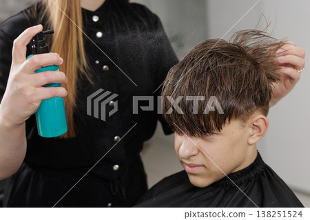 Hairdresser applies styling spray to a boy hair after a haircut. Close-up of hairstyling process, grooming routine, and finishing service in salon. Hairdresser applies styling spray to a boy hair after a haircut. Close-up of hairstyling process, grooming routine, and finishing service in salon. 138251524
