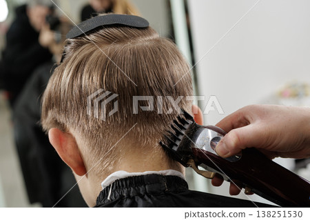 Hairdresser uses an electric clipper to trim the nape area of a boy hair in a barbershop. Close-up of haircut process, grooming service, hygiene, and care concept. 138251530