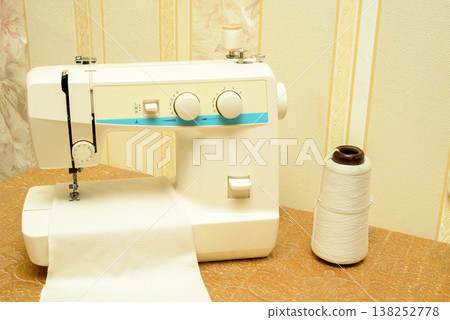 close up of sewing machine with white fabric and large thread cone on handicraft work table surface close up of sewing machine with white fabric and large thread cone on handicraft work table surface 138252778