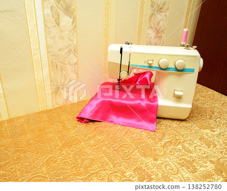 sewing machine on table with bright pink fabric and thread spool against floral wallpaper at home sewing machine on table with bright pink fabric and thread spool against floral wallpaper at home 138252780