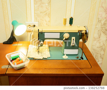 old sewing machine on a table under the light of a green lamp with a thread container nearby old sewing machine on a table under the light of a green lamp with a thread container nearby 138252788