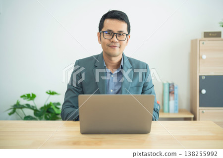 Professional young man in smart casual attire sitting at a desk with a laptop, smiling, representing a modern workspace and remote working environment in a bright room Professional young man in smart casual attire sitting at a desk with a laptop, smiling, representing a modern workspace and remote working environment in a bright room 138255992