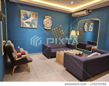 A stylish modern living room interior featuring purple fabric sofas, a unique wavy wooden coffee table, and a large vintage wall clock against deep teal walls with a tall arched gold-framed mirror. A stylish modern living room interior featuring purple fabric sofas, a unique wavy wooden coffee table, and a large vintage wall clock against deep teal walls with a tall arched gold-framed mirror. 138256366