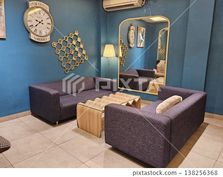 A stylish modern living room interior featuring purple fabric sofas, a unique wavy wooden coffee table, and a large vintage wall clock against deep teal walls with a tall arched gold-framed mirror. A stylish modern living room interior featuring purple fabric sofas, a unique wavy wooden coffee table, and a large vintage wall clock against deep teal walls with a tall arched gold-framed mirror. 138256368