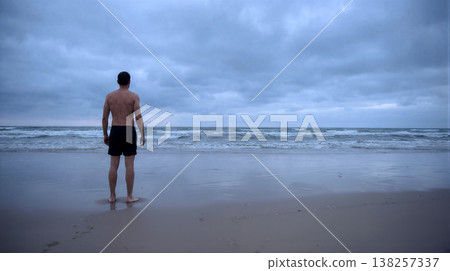 Rear view of shirtless man looking at the ocean on a cloudy beach during twilight 138257337