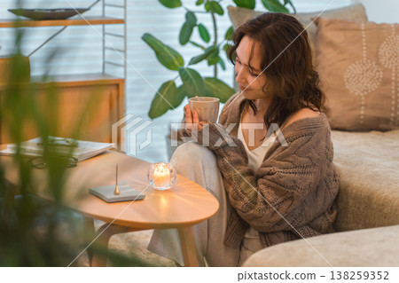 一位時尚的亞洲女性夜晚坐在客廳裡,啜飲著熱飲。 一位時尚的亞洲女性夜晚坐在客廳裡,啜飲著熱飲。 138259352