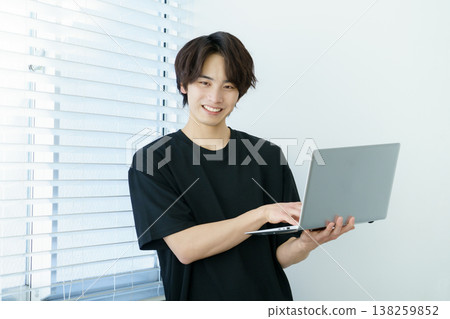一位年輕的日本男子站在窗邊操作筆記型電腦，畫面十分生動。 138259852