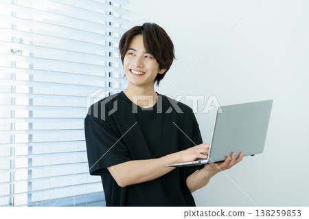 A portrait of a young Japanese man standing by a window and operating a laptop. A portrait of a young Japanese man standing by a window and operating a laptop. 138259853