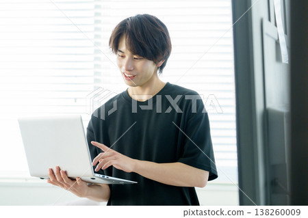 A portrait of a young Japanese man standing by a window and operating a laptop. A portrait of a young Japanese man standing by a window and operating a laptop. 138260009