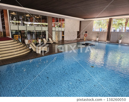 A modern luxury indoor swimming pool featuring crystal clear blue water, elegant wooden decking with sun loungers, and a stylish glass-walled fitness center in a high-end hotel or resort area. A modern luxury indoor swimming pool featuring crystal clear blue water, elegant wooden decking with sun loungers, and a stylish glass-walled fitness center in a high-end hotel or resort area. 138260050
