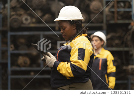 Hispanic female foreman and black female worker standing holding tablet, both of them checking inventory of auto parts and engine maintenance duties in factory. Hispanic female foreman and black female worker standing holding tablet, both of them checking inventory of auto parts and engine maintenance duties in factory. 138261551
