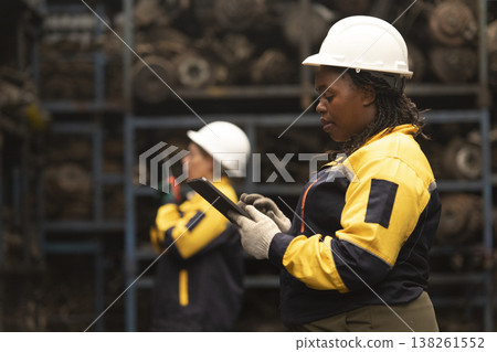 Hispanic female foreman and black female worker standing holding tablet, both of them checking inventory of auto parts and engine maintenance duties in factory. Hispanic female foreman and black female worker standing holding tablet, both of them checking inventory of auto parts and engine maintenance duties in factory. 138261552