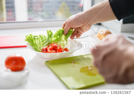 Male Hand Placing Fresh Lettuce Leaves in a Bowl with Sliced Tomatoes for Salad Preparation. Close-up. Male Hand Placing Fresh Lettuce Leaves in a Bowl with Sliced Tomatoes for Salad Preparation. Close-up. 138261567