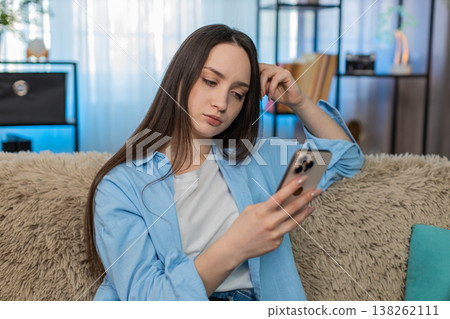 Young woman sitting on sofa shocked receiving unexpected bad news on smartphone sadly holding head 138262111
