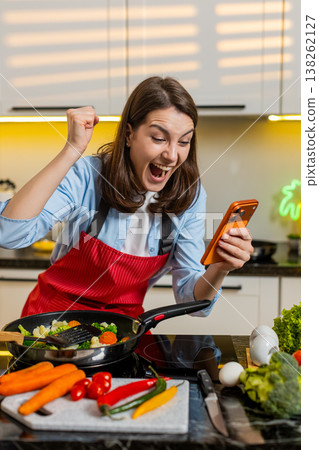 Young woman celebrating prize win on smartphone indoors in modern home kitchen while healthy cooking 138262127