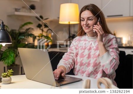 Adult woman talks on smartphone while working on laptop at night voice call smiles productive chat Adult woman talks on smartphone while working on laptop at night voice call smiles productive chat 138262194