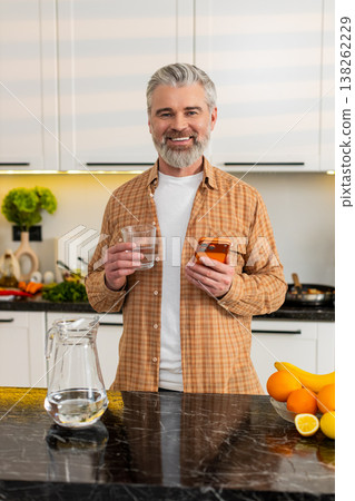 Middle-aged man checks smartphone while drinking water in kitchen for healthy diet hydration routine Middle-aged man checks smartphone while drinking water in kitchen for healthy diet hydration routine 138262229