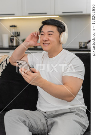 Asian man with wireless headphones enjoys playlist smiles nods to rhythm relaxes with shut eyes calm Asian man with wireless headphones enjoys playlist smiles nods to rhythm relaxes with shut eyes calm 138262656