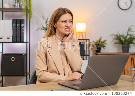 Businesswoman works on laptop winces from tooth pain holds cheek focus keeps typing through bad ache Businesswoman works on laptop winces from tooth pain holds cheek focus keeps typing through bad ache 138263274