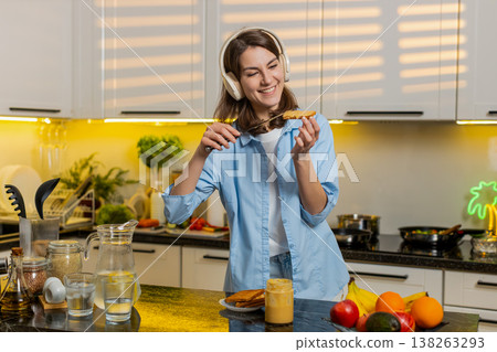 Young woman dancing in home kitchen enjoying peanut butter toast with headphones for diet breakfast 138263293