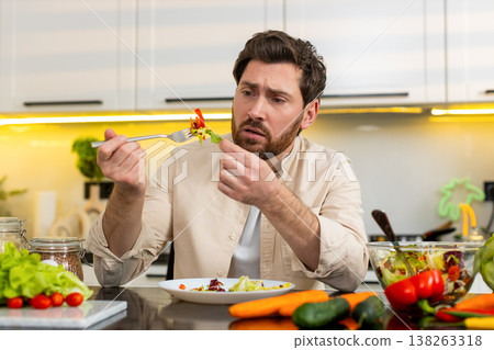 Middle-aged man unhappy eating fresh vegetable salad in home kitchen showing diet willpower struggle 138263318