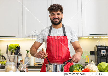 Arabian young adult man smiling stirring soup pot on stove in home kitchen for healthy dinner recipe Arabian young adult man smiling stirring soup pot on stove in home kitchen for healthy dinner recipe 138263319