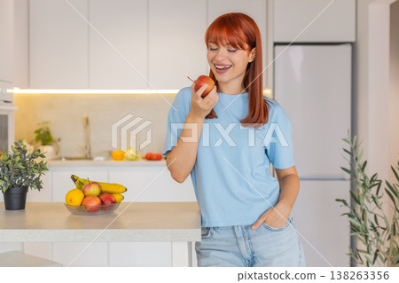 Young woman at kitchen bar takes apple from fruit bowl tosses it up bites and walks away happy smile 138263356
