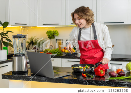 Young woman cooking veggie stir fry while watching laptop recipe video for diet meal in home kitchen Young woman cooking veggie stir fry while watching laptop recipe video for diet meal in home kitchen 138263368