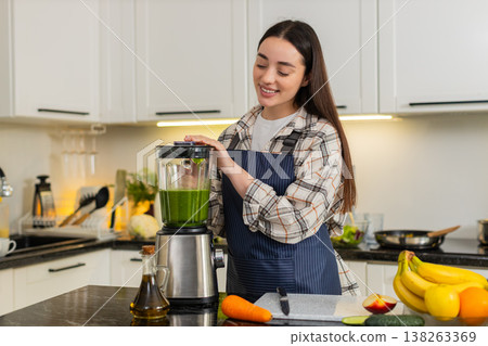 Young woman starts blender for green smoothie with spinach, banana, apple and carrot in kitchen 138263369