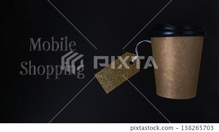 Brown paper coffee cup with black lid and golden tag on dark background, representing mobile shopping and discounts for modern consumers seeking convenience and savings Brown paper coffee cup with black lid and golden tag on dark background, representing mobile shopping and discounts for modern consumers seeking convenience and savings 138265703