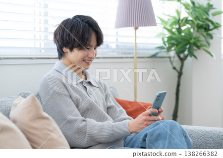 A young man is looking at his smartphone by the living room window. A young man is looking at his smartphone by the living room window. 138266382