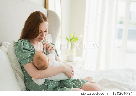 Happy family at home. Mother breastfeeding baby for health nutrition wellness. Mom nursing feeding her newborn child milk at home. Woman infant girl in white bedroom Mother hugs baby with love care 138266784