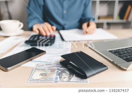 Tax budgeting workspace with cash, wallet, calculator, smartphone and laptop on office desk, while accountant writes notes on paperwork, supporting income filing, deductions, compliance and audit. Tax budgeting workspace with cash, wallet, calculator, smartphone and laptop on office desk, while accountant writes notes on paperwork, supporting income filing, deductions, compliance and audit. 138267437