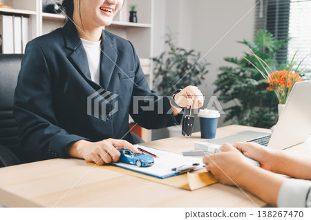 Smiling agent holding car key and pointing to blue model car beside contract while client counts cash at office desk with laptop, symbolizing auto purchase, payment, dealership service and trust. Smiling agent holding car key and pointing to blue model car beside contract while client counts cash at office desk with laptop, symbolizing auto purchase, payment, dealership service and trust. 138267470