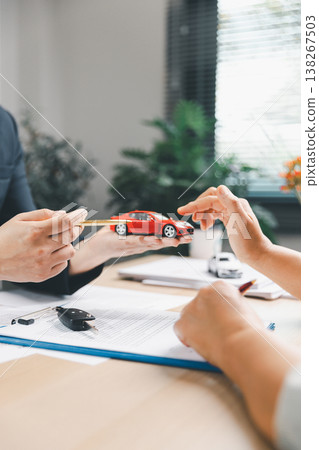 Agent presenting red model car and pen to client above contract with car key on office desk, representing vehicle purchase consultation, ownership planning, dealership service and trust. Agent presenting red model car and pen to client above contract with car key on office desk, representing vehicle purchase consultation, ownership planning, dealership service and trust. 138267503