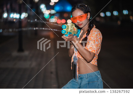 Confident young woman aiming water gun at Songkran night celebration, wearing bright orange goggles on an urban street with colorful bokeh lights, expressing playful attitude, festival fashion and 138267592