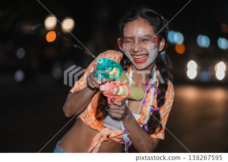 Excited woman winking and aiming two water guns during Songkran night festival, with face powder, safety goggles and glowing street bokeh, expressing playful action, summer party mood and youthful 138267595