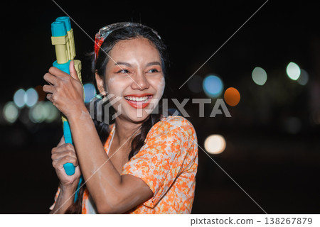 Portrait of smiling young woman holding water gun during Songkran night celebration, with wet face, soft bokeh lights and festive styling, showing cheerful Thai holiday mood, street fun and summer Portrait of smiling young woman holding water gun during Songkran night celebration, with wet face, soft bokeh lights and festive styling, showing cheerful Thai holiday mood, street fun and summer 138267879