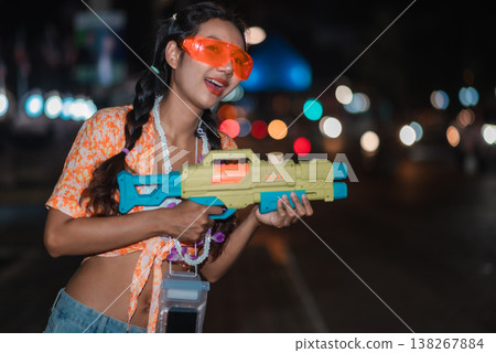 Stylish woman carrying water gun during Songkran nightlife celebration, wearing orange goggles and floral shirt on a city street with blurred lights, capturing trendy festival portrait, summer fun and 138267884